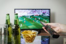 The Best Time to Advertise on TV Blog Cover - Hand holding TV remote pointed at sporting event with drinks and a cup of chips in the foreground
