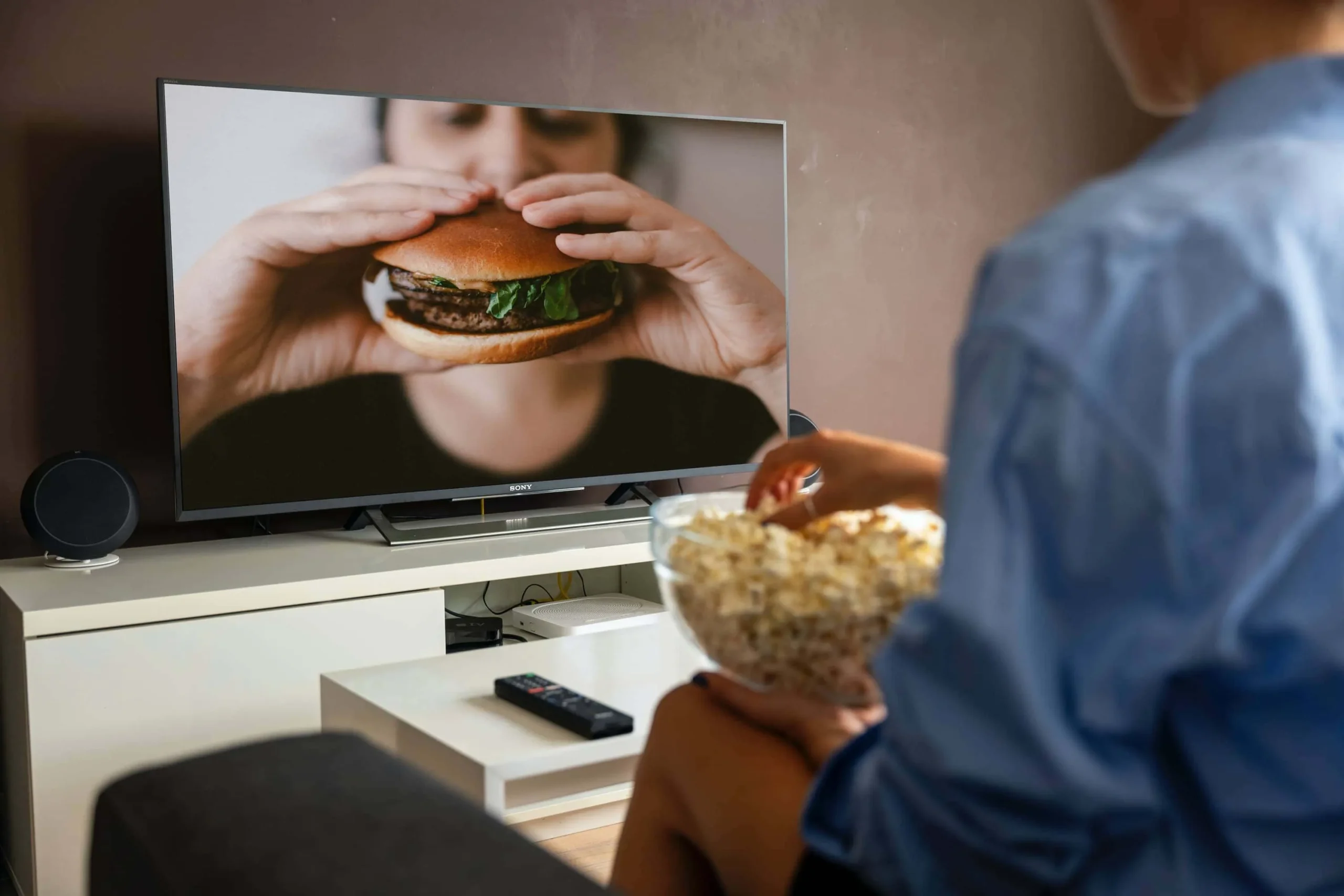 Man sitting on couch with a bowl of popcorn watching a burger commercial on TV 